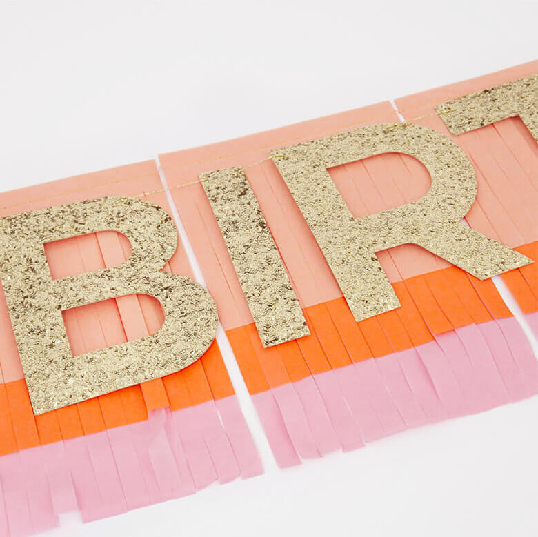 close up details of Meri Meri Pink Happy Birthday Fringe Garland. There are two layers of tissue paper are folded together before being cut into fringing ,The fringed pennants are made of pink, peach and coral tissue paper, The glittered letters are pre-threaded onto cord, and the fringing is stitched together with peach satin ribbons