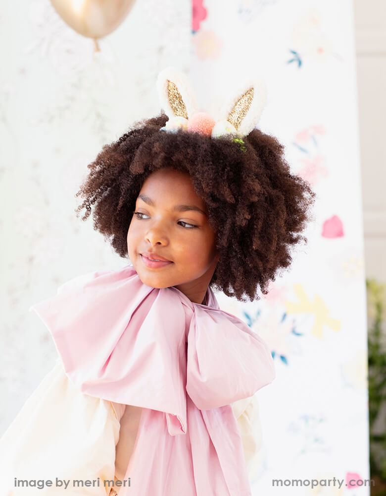 A little african american girl wearing a pink dress and a Meri Meri Pom Pom Bunny Ear headband with glittery ears and lots of colorful pompoms on her curly hair, for a cutest modern Easter celebration