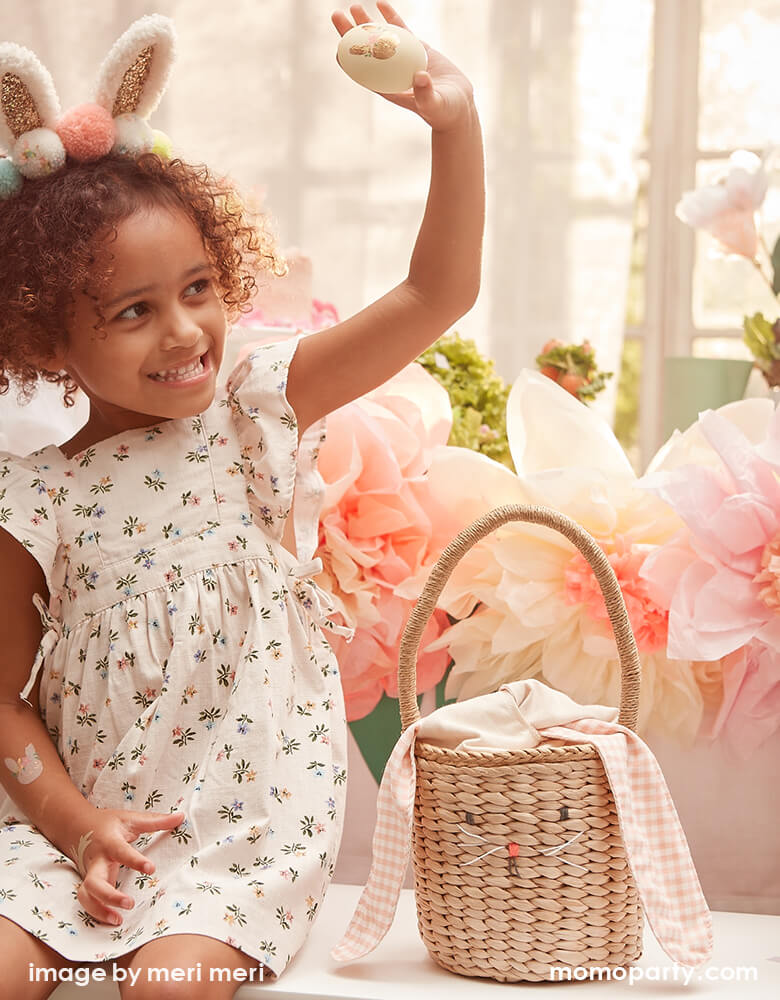 A girl wearing a Meri Meri Pom Pom Bunny Ear headband with glittery ears and lots of colorful pompoms, holding a egg decorated with egg stickers, with her bunny easter basket on the side sitting in a room decorated with floral paper garland for the cutest modern Easter egg hunt