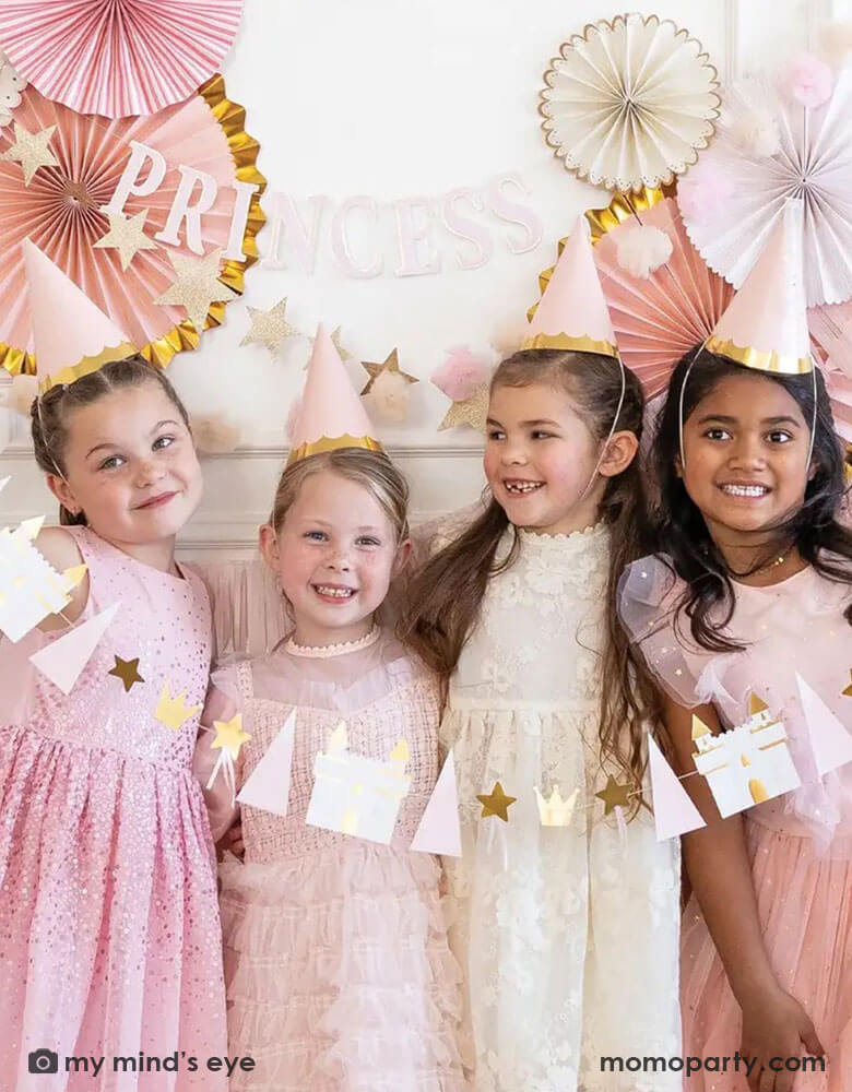 A group of 4 girls in their 6 or 7 years old dressed up as princesses with their tutu dresses and Momo Party's princess birthday hats, they're standing in front of a wall decorated with pink party fans with gold foil. Together they hold Momo Party's 5.5 ft princess icon banner featuring castles, crowns, and stars. A sweet look for girl's princess themed birthday party.