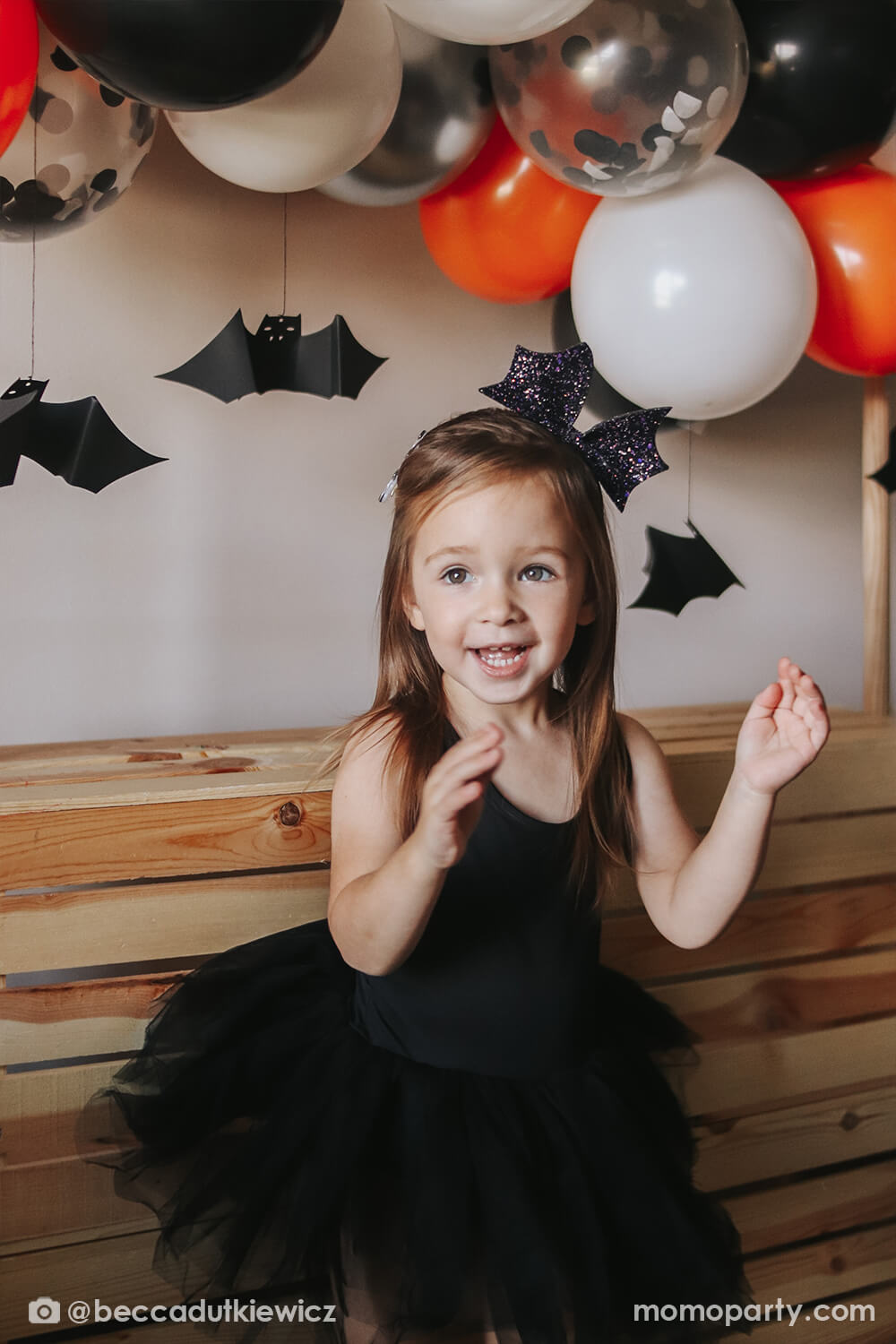 Girl wearing black tutu dress and Bat Bow Headband by Great Pretenders, with Meri Meri Halloween Balloon Garland Kit in a orange, black white, with confetti latex balloon and hanging bat decoration on the back. This sparkly bat bow headband in purple color will add an extra Halloween flair to any outfit! Beautiful and affordable Accessory for girl, Accessory for kid sold by party boutique online store momoparty.com