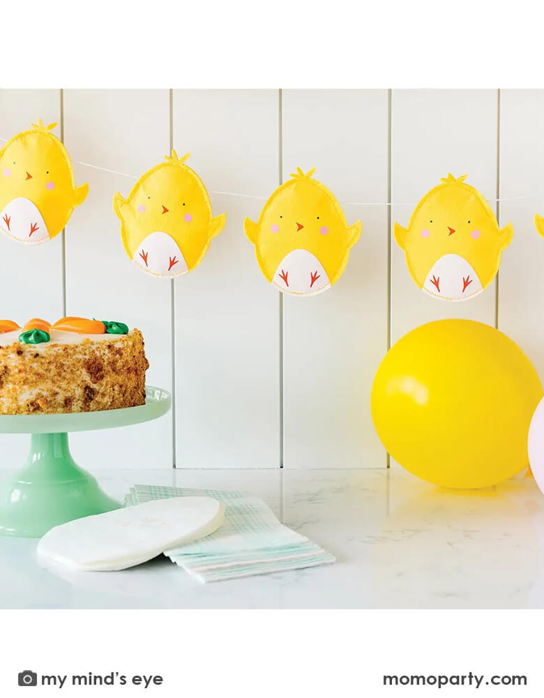 A kitchen counter decorated with Momo Party's 6 ft puffy felt chick banner by My Mind's Eye, with a carrot cake and easter egg shaped napkins and blue gingham guest towels, it creates a simply yet unique look for an Easter celebration.