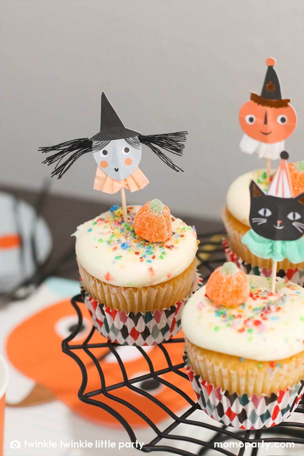 Kid's Halloween party table filled with Halloween themed party supplies including a set of cupcakes decorated with Halloween character toppers including a black cat, a skull, a pumpkin, and a witch on a spider cobweb shaped cupcake stand, creating a spooky vibe for this Halloween season!