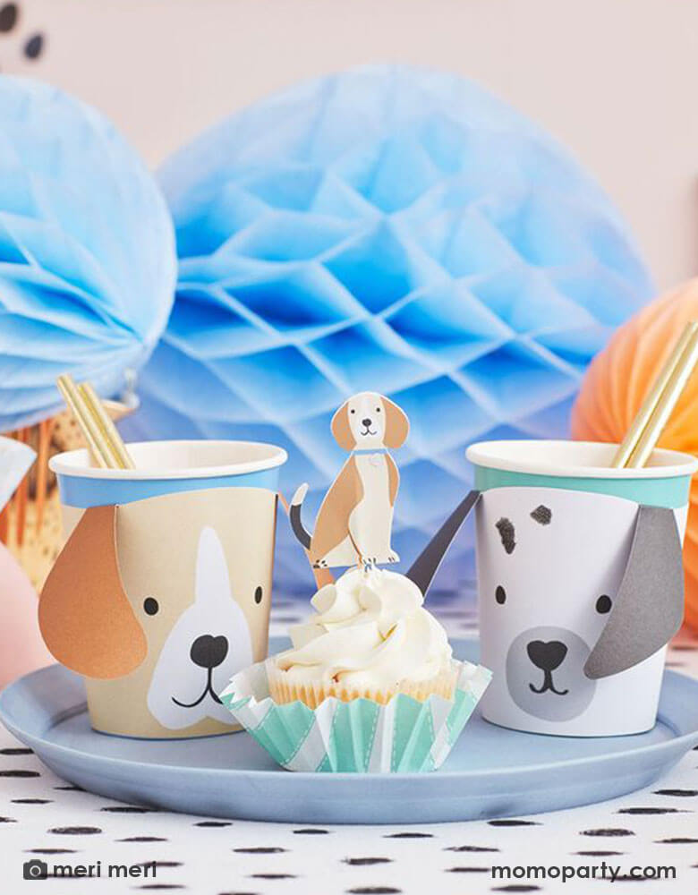 A party table featuring adorable dog themed party goods including Momo Party's 9oz puppy party cups by Meri Meri and cupcakes decorated with Meri Meri's puppy cupcake toppers. In the back it's a bunch of blue and peach colored honeycomb decorations, all together makes a great inspiration for a modern and chic dog themed party for kids or dog-loving adults.