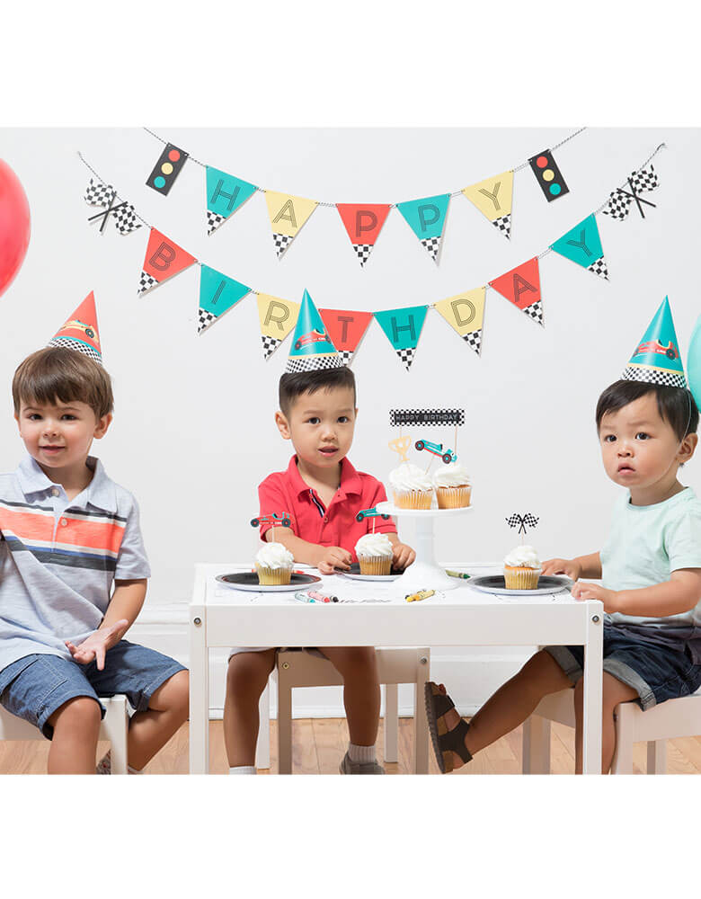 A Race car themed birthday party with three boys featuring Merrilulu Race Car Happy Birthday Banner Set on the wall with color flags and checkered race car flag designs. On the party table there's cupcakes topped with merrilulu's race car cake toppers