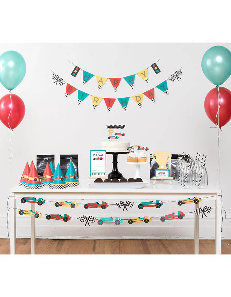 A Race car themed birthday party set up with Merrilulu's Race Car party collection including a Happy Birthday Banner Set  with color flags and checkered race car flag designs on the wall, party hats with race car illustrations,  cupcakes topped with race car cake toppers on the party table. under the table there's a fun colorful race car garland set hung to add some festivities to the party