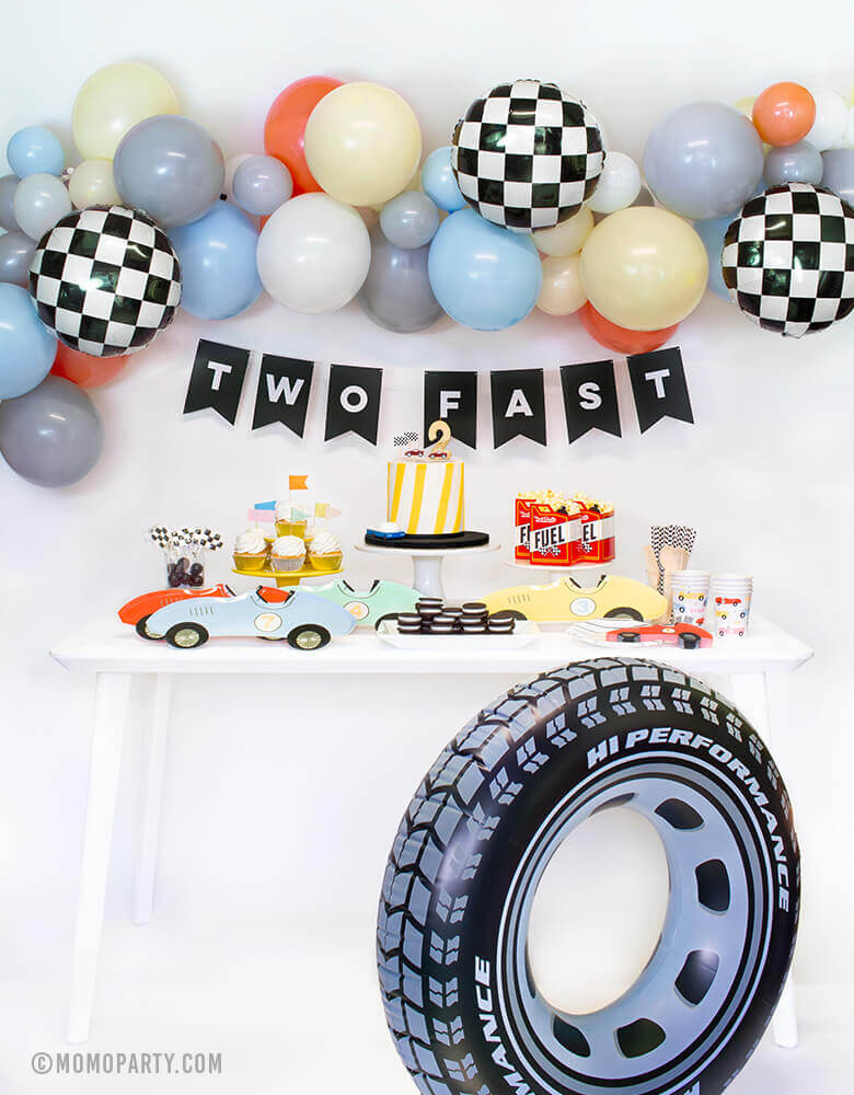 Momo Party Race Car Box, set up inspiration with Meri Meri race car paper plates, race car shape napkins, race car graphic party paper cups, black strip small plate, neon confetti flag cupcake kit, race Car candles and gold #2 candle over a cake, Fuel treat favor box with popcorns as tableware, colorful balloon cloud, black costume letter banner with script of "Two Fast", checkerboard foil balloon and Inflatable giant tire tube as decorations, for a 2 years old modern Race car, car themed birthday party