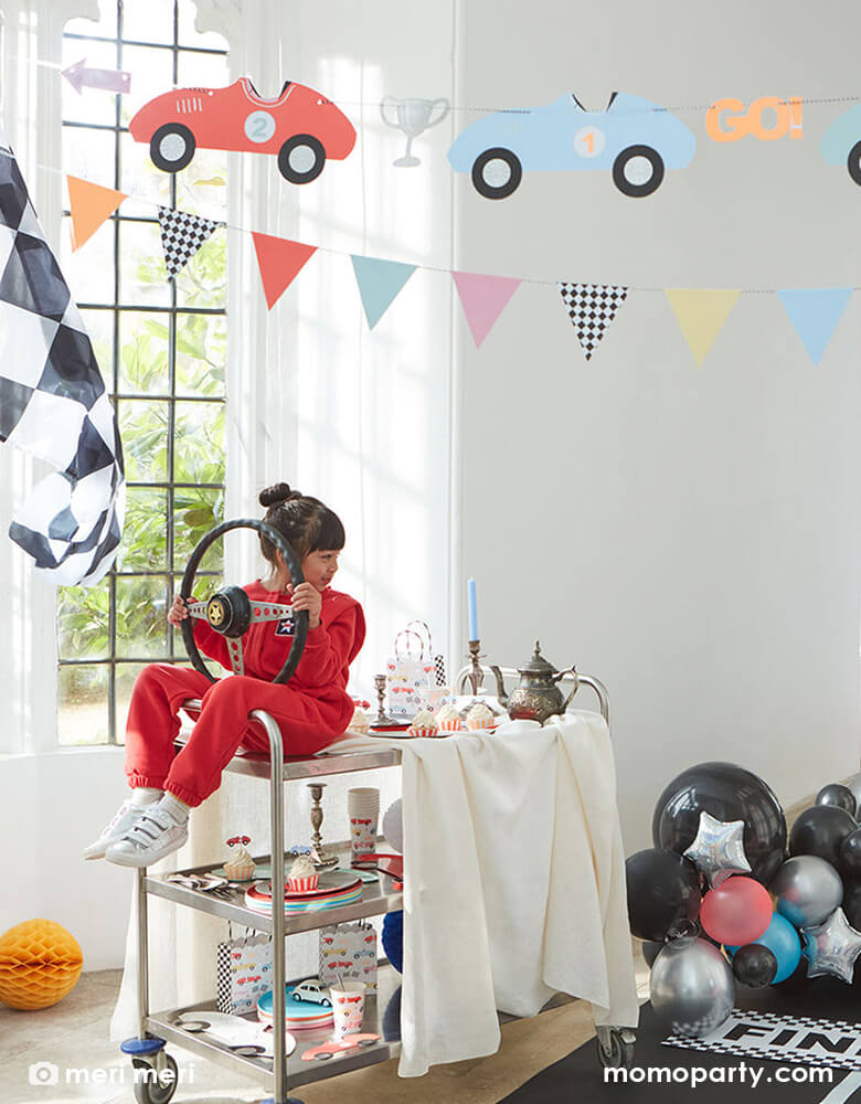 Race car party in the house, decorated with Meri Meri Race Cars Garland,  balloons, race car checker flags, with a birthday girl wearing a red racing suit, holding a car Steering Wheel pretending like driving by sitting on a cart with all the car themed party tableware