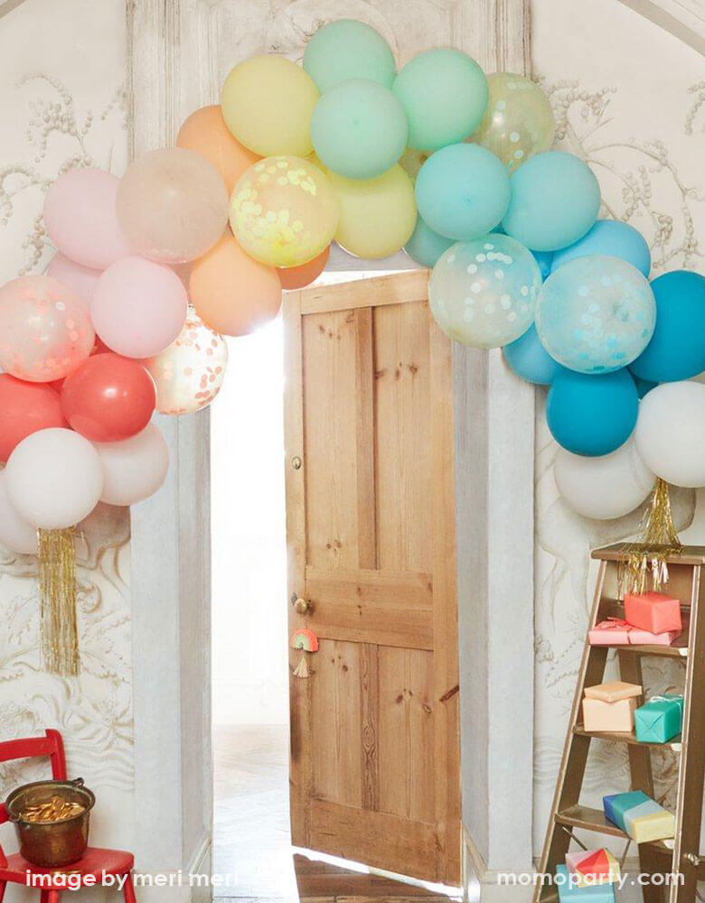 A room decorated with Meri Meri's Rainbow Balloon garland Arch Kit with multicolored balloons, confetti balloons, and gold tinsel  hung over the door of the room