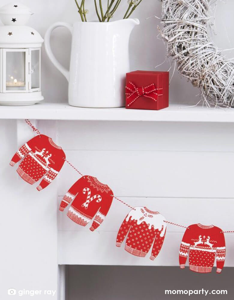 Mantelpiece decorating with Ginger Ray - RED AND WHITE FESTIVE JUMPER WOODEN BUNTING - COSY CHRISTMAS. And there is white jar and white candle house and small red gift box on top of it. This Red and White Christmas Sweater Wooden Garland featuring 8 sweater pennant in red and white color. Create a cozy feel in your home this Christmas with this unique red and white Christmas Sweater wooden garland. Hanging on walls for your holiday celebration and ugly sweater party
