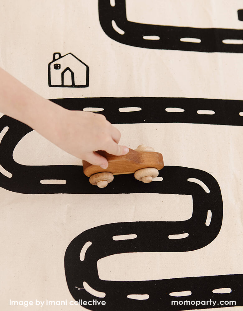 A close up look of A Boy's hand holding a wooden car toy on Imani Collective interactive play floor mat - Road Floor Mat. This floor mat is natural canvas hand with black screen printed road bike, houses modern illustration, Sewn and screen printed by hand on natural canvas by Kenyan artisans. Sold by Momo party store provided modern party supplies, boutique party supplies