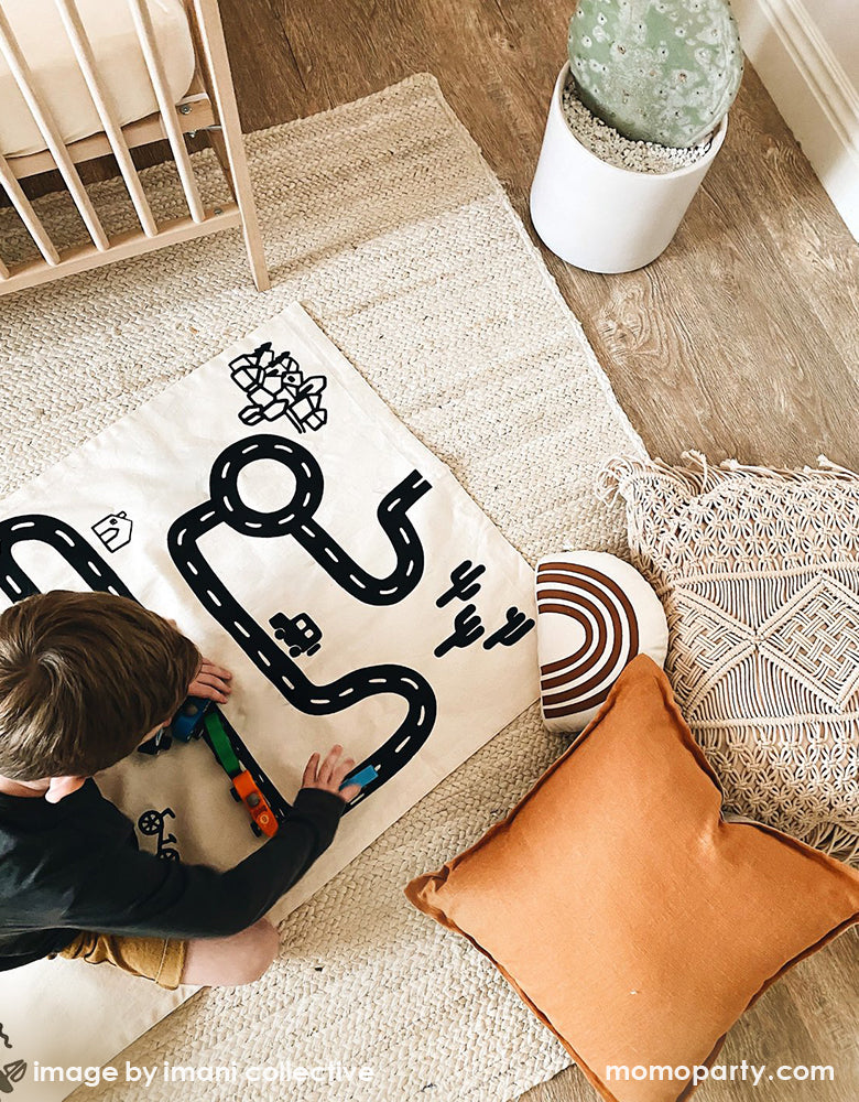 A top view of a  Boy play with his wooden car toys on Imani Collective interactive play floor mat - Road Floor Mat in his modern playroom. This floor mat is natural canvas hand screen printed, Sewn and screen printed by hand on natural canvas by Kenyan artisans. Sold by Momo party store provided modern party supplies, boutique party supplies