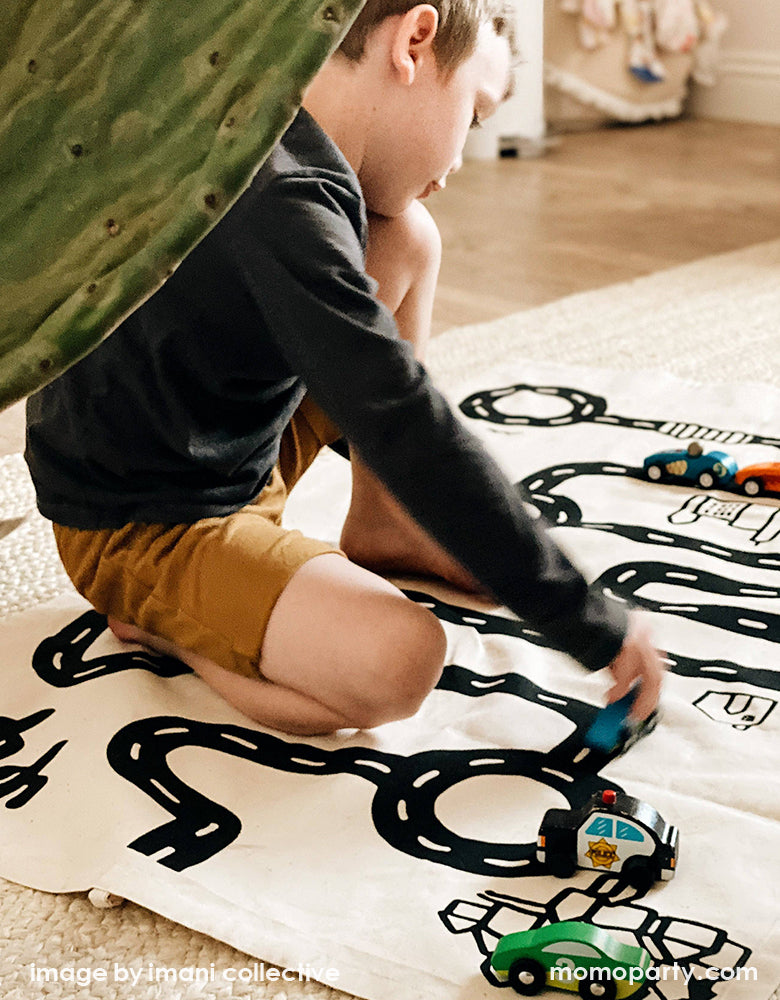 A Boy play with his wooden car toys on Imani Collective interactive play floor mat - Road Floor Mat in his modern playroom. This floor mat is natural canvas hand screen printed, Sewn and screen printed by hand on natural canvas by Kenyan artisans. Sold by Momo party store provided modern party supplies, boutique party supplies