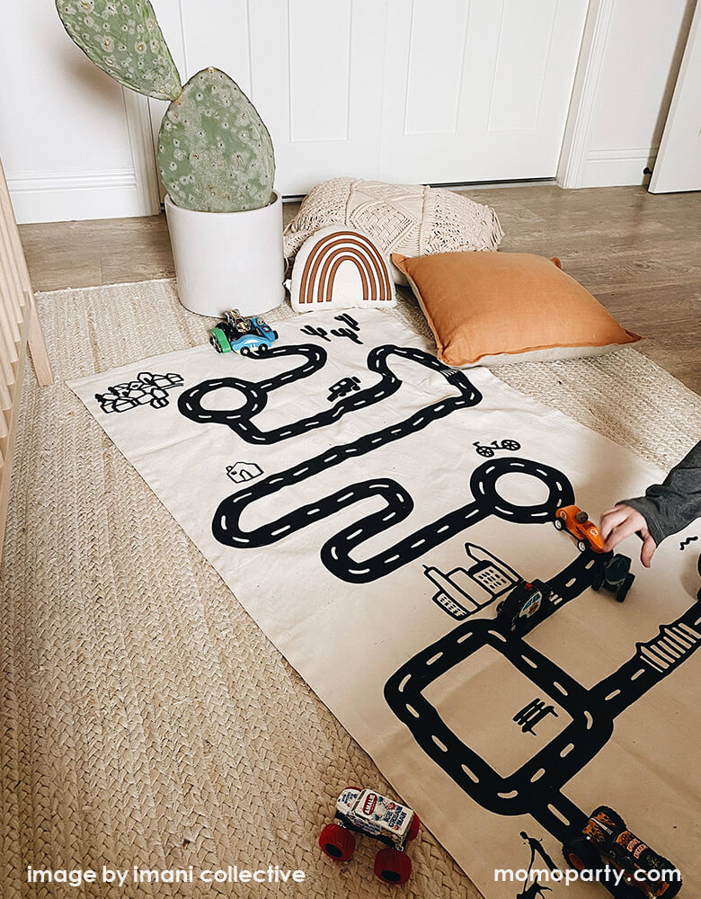 A kid play with his wooden car toys on Imani Collective interactive play floor mat - Road Floor Mat in his modern playroom with rainbow pillow, plants around. This floor mat is natural canvas hand screen printed, Sewn and screen printed by hand on natural canvas by Kenyan artisans. Sold by Momo party store provided modern party supplies, boutique party supplies