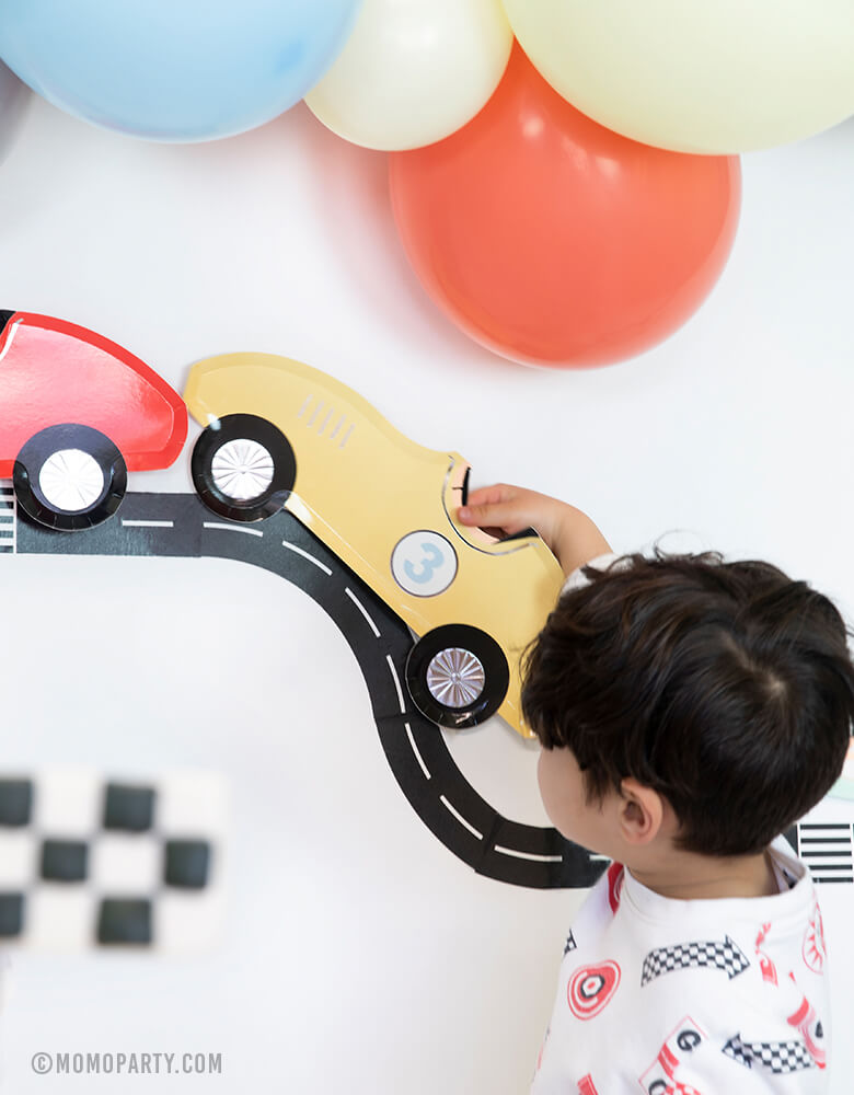 boy play with MeriMeri Car Plate with Road tape decorated as backdrop with matt blue, coral, matt yellow balloon garland in a race car birthday party