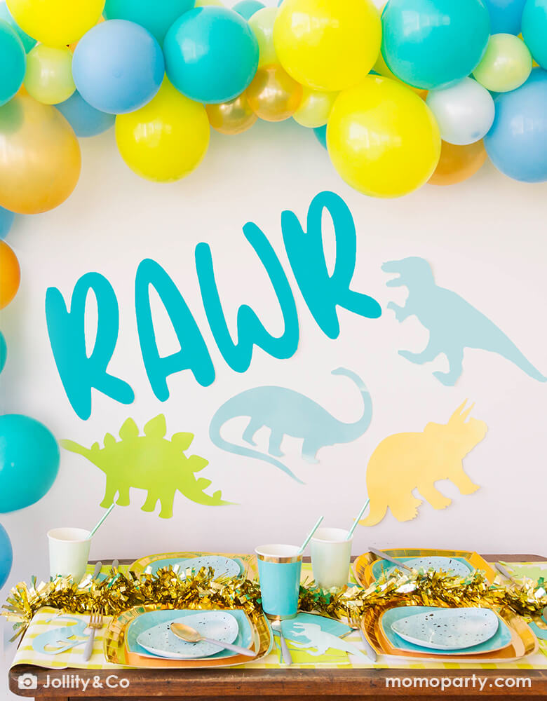 Dinosaur themed party set up with yellow, caribbean blue and pale blue colored balloon garland on the wall, a RAWR sticker with dinosaurs shaped paper cuts as backdrop,  Rockin' Robin Dinner Plates layered with Posh Chill Out Plates, some dinosaur shaped paper cuts, on a yellow gingham table runner. for a unique fun dinomite birthday party