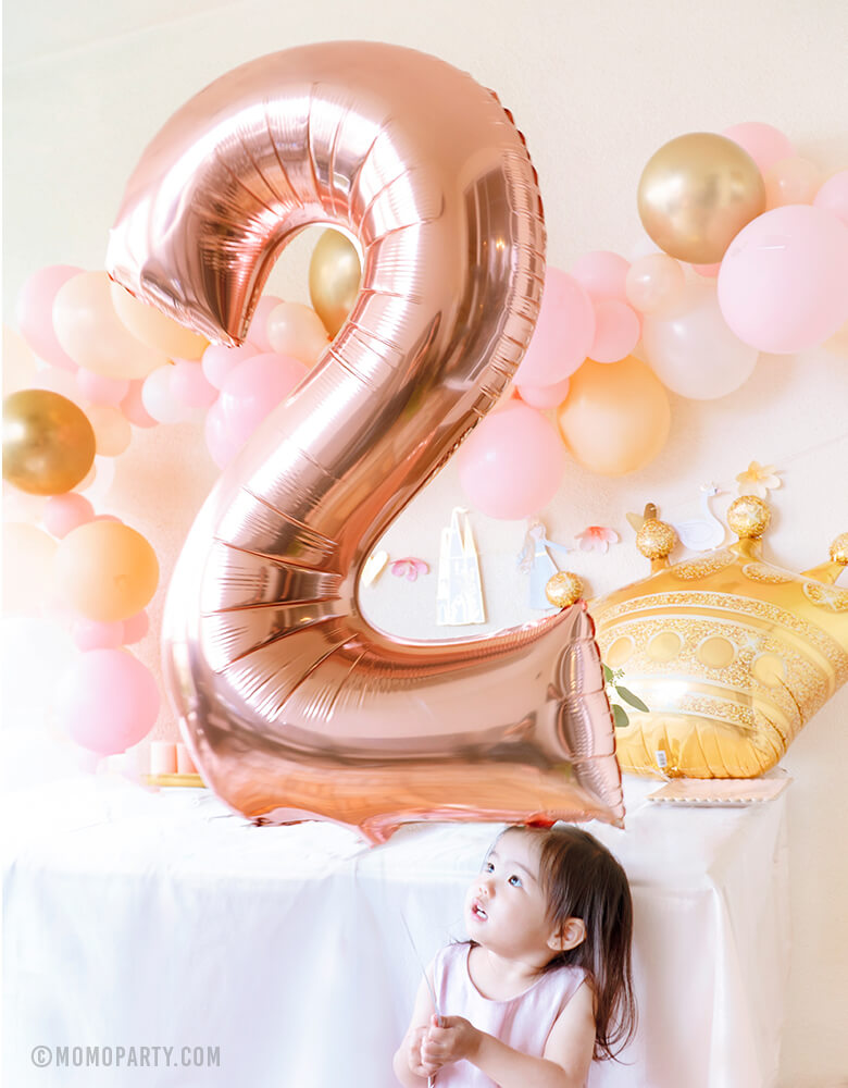 Little princess holding a Large Number Two Rose Gold Foil Mylar Balloon, with a Gold Crown foil balloon, pink gold colored balloon garland on the back for her 2nd princess themed birthday celebration.