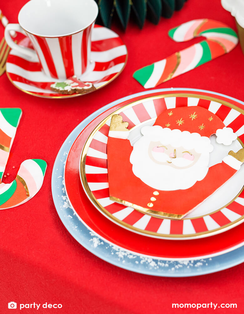 Holiday table with Santa Napkins layered on a red striped small plate, medium red plate and a large light blue plate. a Candy cane Napkins mixed with red pink green and gold colors, a red stripe tea set, gold utisles on a red tablecloth. These jolly Santa napkins printed with red white and gold foil color with raising arms and happy face, there are gold stars on his hat. these cute holiday party supplies will add festive cheer to your Christmas celebration with the little ones!