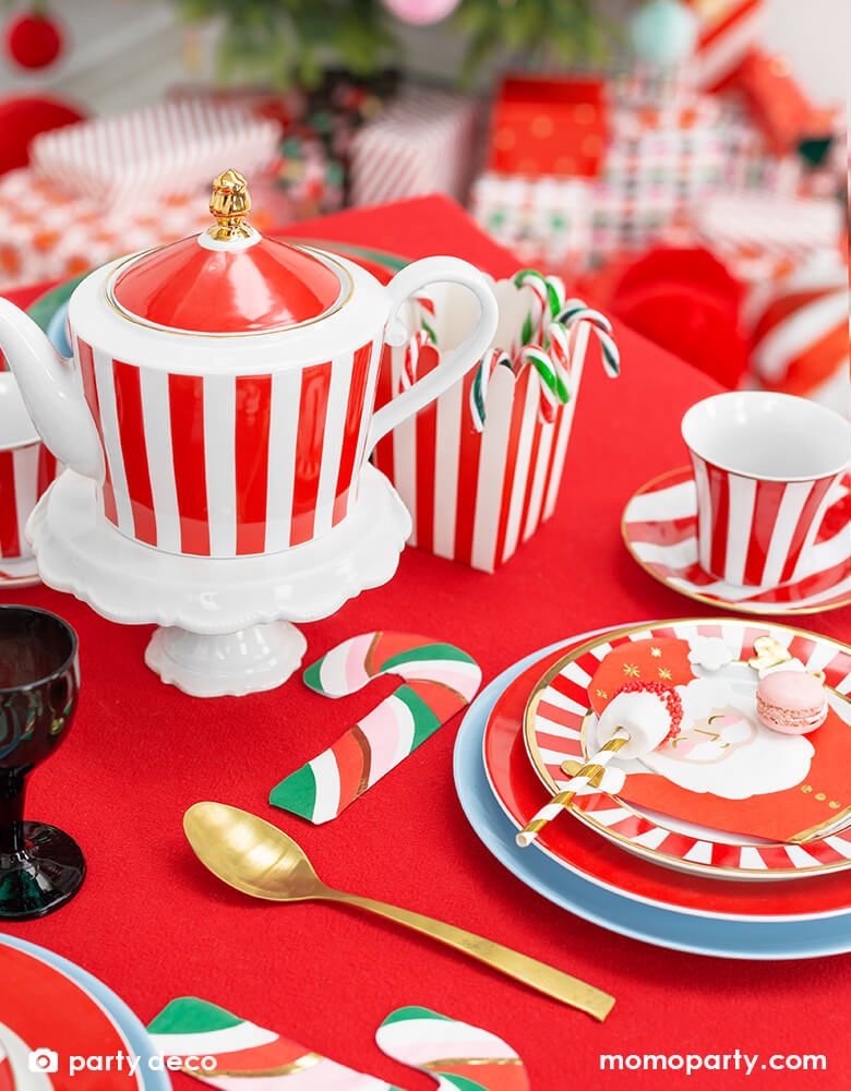 Holiday table with Santa Napkins layered on a red striped small plate, medium red plate and a large light blue plate. a Candy cane Napkins mixed with red pink green and gold colors, candy cane in a red stripe popcorn box, a red stripe tea set, gold utisles all on a red tablecloth. The jolly Santa napkins with raising arms and happy face, there are gold stars on his hat. these cute holiday party supplies will add festive cheer to your Christmas celebration with the little ones!