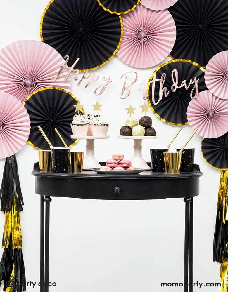 A pink and black and gold themed birthday party set up with paper fans on the wall and an elegant dessert table for pink cupcakes topped with gold star toppers, a great party look for a girl's Paris themed birthday party