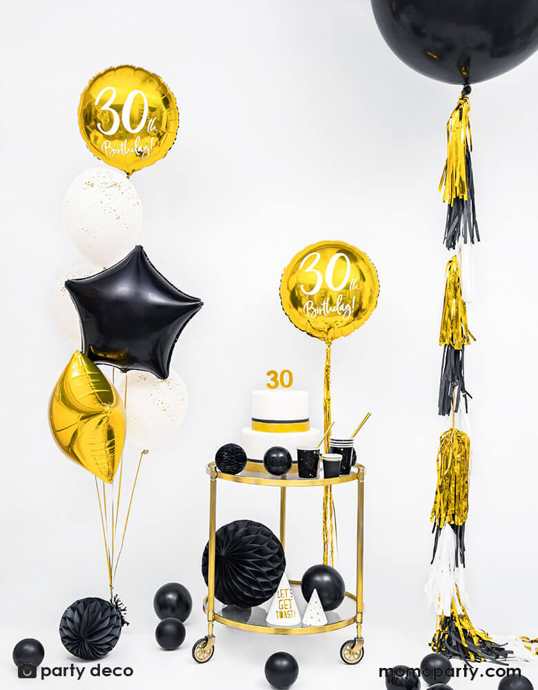 A gold and black themed 30th birthday party featuring black jumbo balloon with gold and black tassel, along with a bar cart with a birthday cake on top