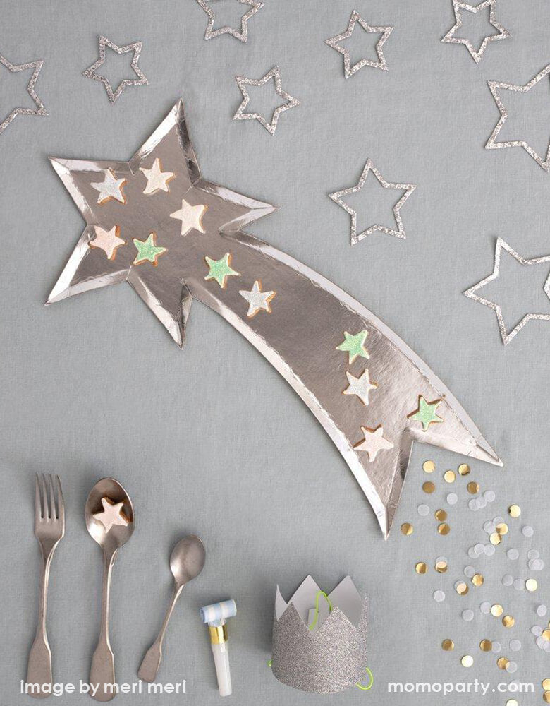 Top Party table view with small star shaped cookies on Meri Meri Shooting Star Platters, a set of silver utensils, Silver Sparkle star decorations, Bright Stripe Party Blowers, and Mini Silver Glitter Crowns and confettis on a grey tablecloth . These shooting star die cut shaped paper party platters are beautifully designed and crafted with a shiny silver foil finish, they are perfect as a table centerpiece or for passing canapes around on.