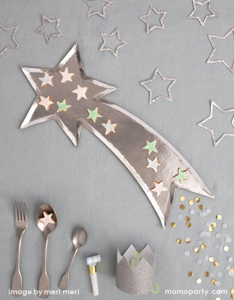 Top Party table view with small star shaped cookies on Meri Meri Shooting Star Platters, a set of silver utensils, Silver Sparkle star decorations, Bright Stripe Party Blowers, and Mini Silver Glitter Crowns and confettis on a grey tablecloth . These shooting star die cut shaped paper party platters are beautifully designed and crafted with a shiny silver foil finish, they are perfect as a table centerpiece or for passing canapes around on.
