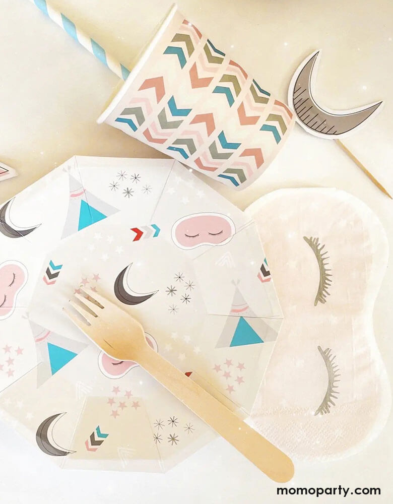 A tablescape of girls sleepover party featuring Momo Party's sleepover napkins in the shape of sleeping masks by Pooka Party, along with coordinated plates featuring  sleepover themed illustrations like sleeping tents, sleeping masks, moons and stars, plus light pink party cups with chevron design, and cupcake topers with moon design, perfect for girl's slumber party.