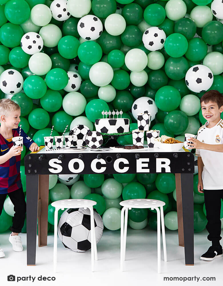 two boys celebrating birthday in front of a balloon wall with different shades of green balloons and soccer ball balloons, with a party table with soccer themed party supplies and tableware, perfect ideas for kid's soccer themed birthday party!