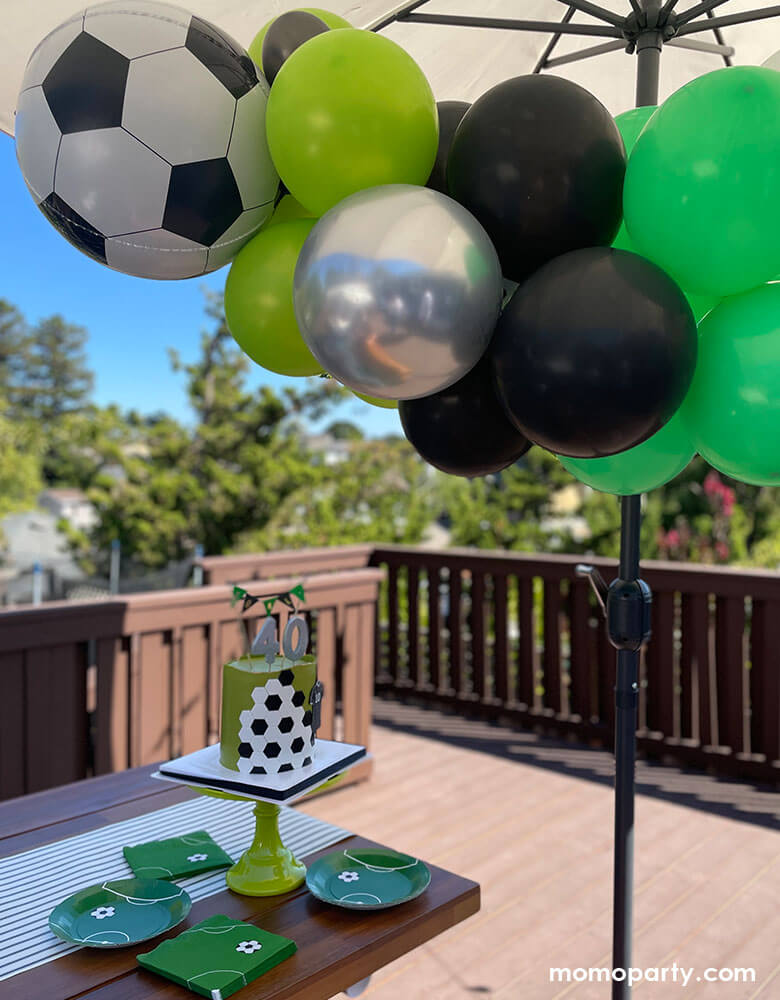 A backyard soccer themed birthday party with a soccer balloon garland hung under the patio umbrella and soccer themed plates and napkins in green with modern soccer design, next to them is a soccer themed birthday cake topper with silver number birthday candles and "goal" cake topper in black and green with gold detail.