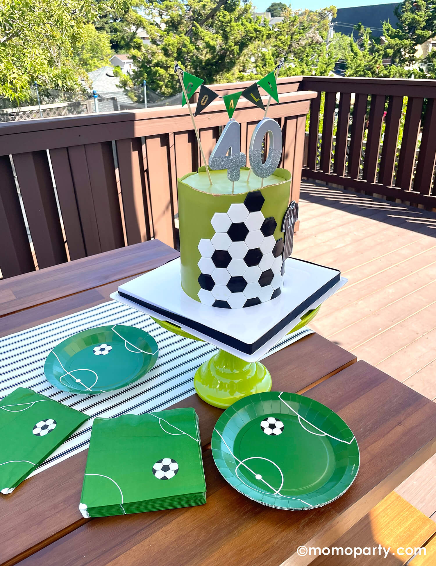 A backyard soccer themed birthday party with soccer themed plates and napkins in green with modern soccer design, next to them is a soccer themed birthday cake topper with silver number birthday candles and "goal" cake topper in black and green with gold detail.