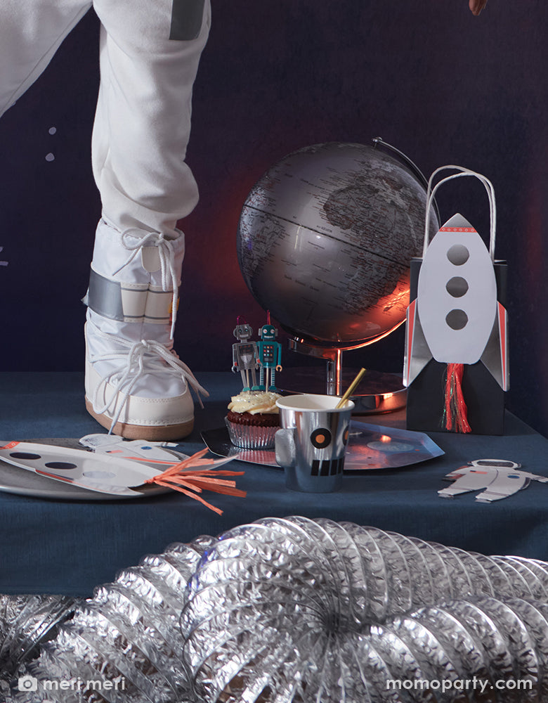 A fun party table with Meri Meri's space themed party tableware including some space dinner plates with planets design, an astronaut shaped napkins with silver foil, a spaceship shaped large plates with silver foil accent and tassel decorations, along with space robot party cups, with a kid's foot in a space suit and globe in the background - a whimsical look and inspiration of kid's space themed birthday party decorations