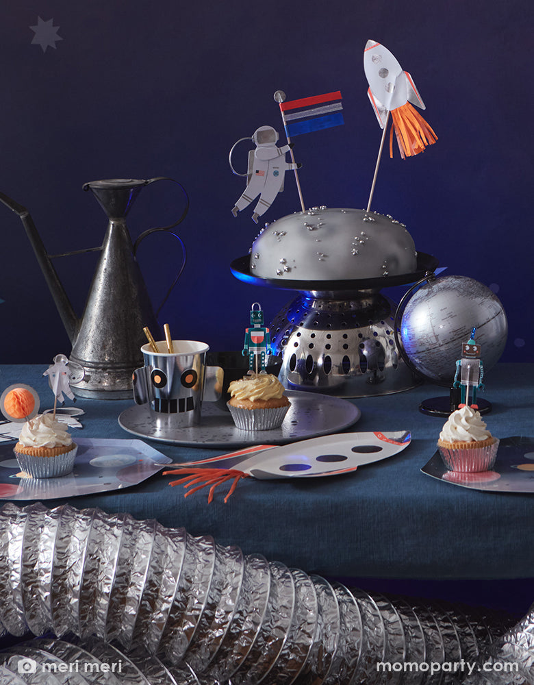 Close up of space themed party table with Rocket Plates by Meri Meri, cupcakes decorated with space cupcake toppers on a Space Dinner Plate, Robot Cup with gold straws, silver moon cake set decorated with cake toppers, a silver globe. These modern unique designed party supplies are perfect for a space themed birthday party, blast off birthday party, two the moon birthday party.