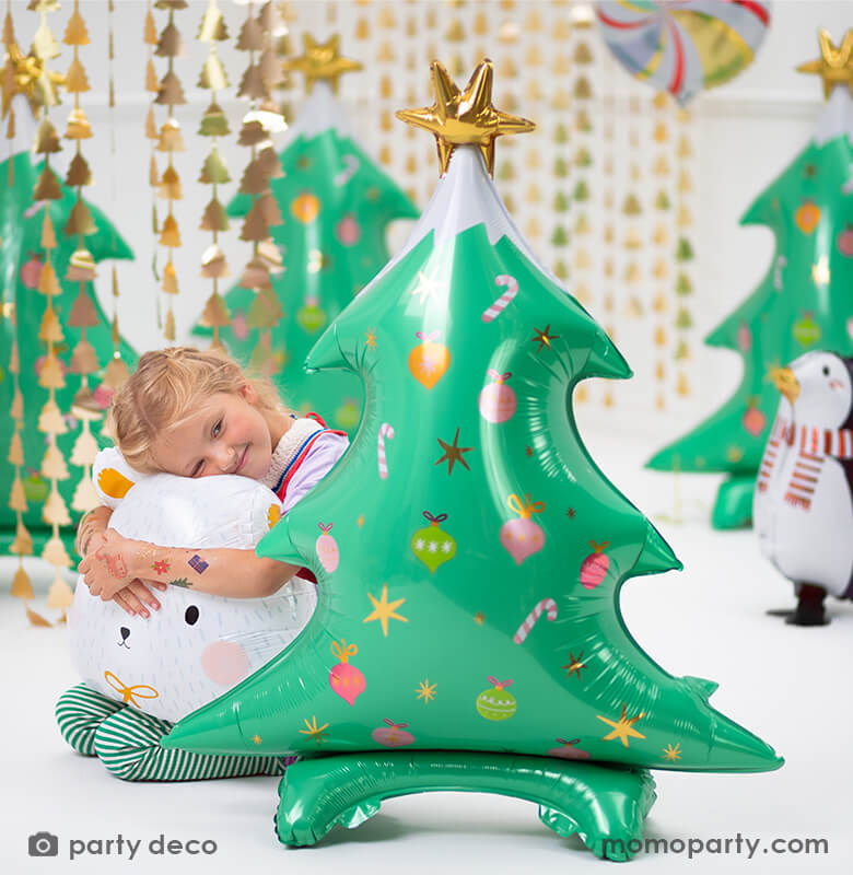 A sweet girl holding a polar bear foil balloon, hiding behind a Party Deco - Standing Christmas Tree Foil Balloon. This 37 inches adorable standing Christmas tree in matte finishing with gold satin print, a gold star on the top of the tree, with candy cane, ornaments graphic print, in a room decorated with Christmas Tree Gold Backdrop, and lot of christmas themed foil balloons, These modern and unique holiday party supplies will delight your Holiday celebration.