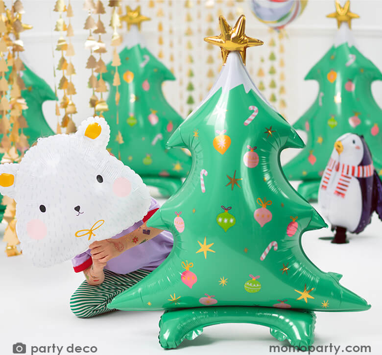 A sweet girl covering her face with a polar bear foil balloon, peeking behind a Party Deco - Standing Christmas Tree Foil Balloon. This 37 inches adorable standing Christmas tree in matte finishing with gold satin print, a gold star on the top of the tree, with candy cane, ornaments graphic print, in a room decorated with Christmas Tree Gold Backdrop, and lot of christmas themed foil balloons, These modern and unique holiday party supplies will delight your Holiday celebration.