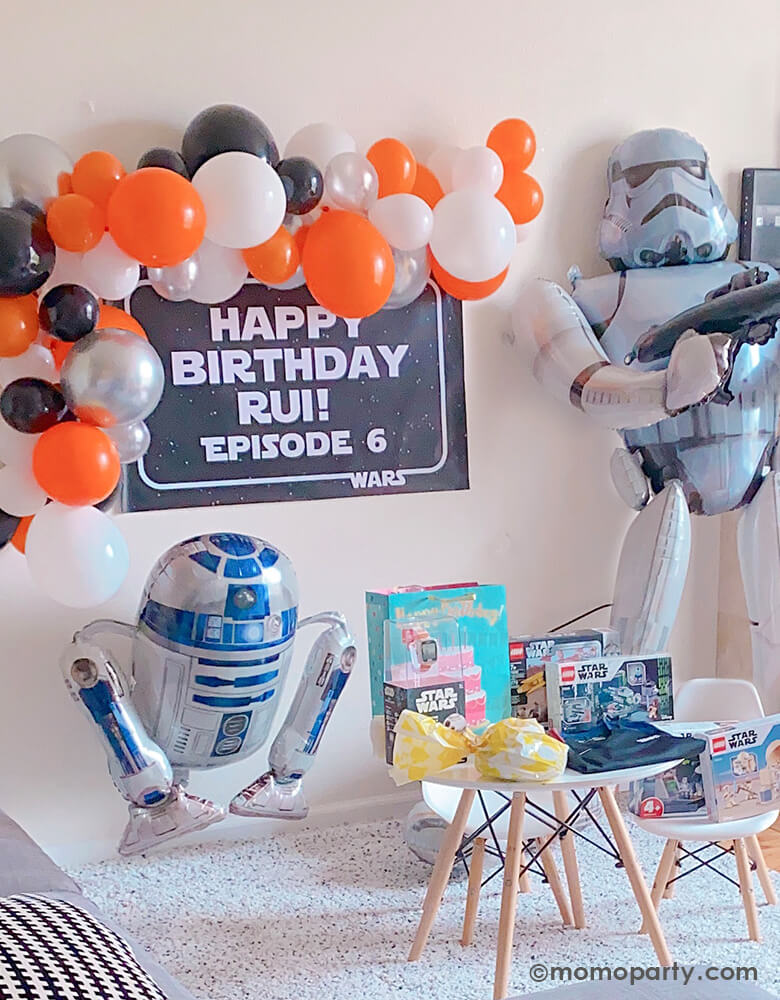 Boy's 6 years Star War themed birthday decorated with a orange white and black themed balloon garland, a black banner with "happy birthday Rui! Episod 6" in Star war typeface, a Star Wars R2D2 Foil Balloon stand on the bottom, and a Giant Stormtrooper AirWalker Foil Balloon stand on the side, and lots of star war themed birthday gift on the kid's desk, ready to celebration his star war birthday