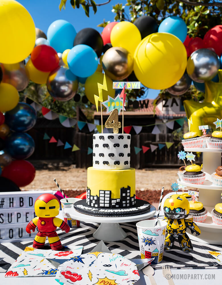 Kids modern superhero birthday party table set up with Day Dream Society Superhero Party Small Paper Plate, Star Silver Foil Plate, Superhero Napkins,superhero cup and wooden utensil, Modern Superhero cake with number 4 gold gilter candle, superhero sign cake topper, superhero figure toys, balloon garland as decoration on the back