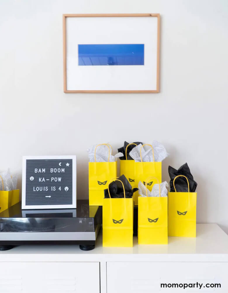 A table with yellow goodie bags decorated with Momo Party's superhero mask stamp, on the left is a modern letterboard spells out Bam Boom Ka-pow for a boy's birthday party celebration