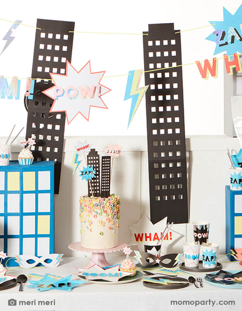 Superhero themed birthday party table, filled up with superhero comic word shaped plates, superhero cups, neon Lightning Bolt Napkins, a colorful chorios cake decorated with superhero cake toppers, cupcakes with superhero cupcake toppers, superhero party masks. And a large superhero garland with fun statements "Boom!, Pow, Wham! and Zap!" and Black towering skyscrapers with cut out windows hanging on the wall. Such a fun, bold, easy set up and  modern superhero themed birthday for any superhero lovers