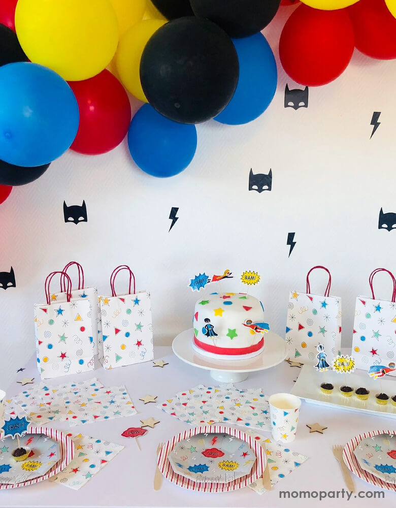 Modern superhero birthday party decoration ideas, with blue, red, yellow, and black color mixed balloon garland hanging, with the wall decorate with pooka Superhero Reusable Wall Decal in a superhero masks and lightning bolts black vinyl stickers, and colorful superhero themed party table wares on the table, a cake in the middle with superhero elements, and Happy Colors Party Bags next to it. These modern and fun superhero themed party supplies are perfect for your superhero's celebration.