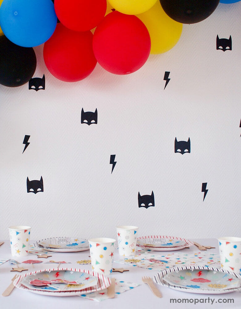 Modern superhero birthday party decoration ideas, with blue, red, yellow, and black color mixed balloon garland hanging, with the wall decorate with pooka Superhero Reusable Wall Decal in a superhero masks and lightning bolts black vinyl stickers, and colorful superhero themed party table wares on the table. Simple and fun party supplies for your superhero's celebration.