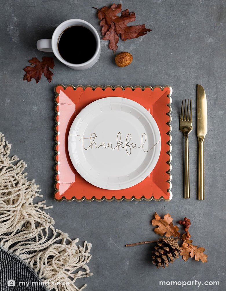 Table top view of My mind's Eye 7 inches Thankful Grateful Plates with "thankful" printed in a beautiful script in gold foil on the cream paper plates, layered with Orange with golden foil scalloped large plates, there are golden cutlery, a cup of coffee, some dry Maple leave, nuts around on top of the  grey quartz countertop. These modern party supplies are Perfect for your Thanksgiving celebration dessert or appetizers. 