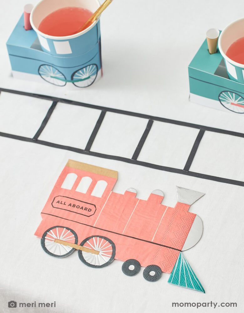 A train themed party table with train track decoration and Meri Meri's train party cups and red train shaped napkin, crafted in 3-ply paper with silver foil in a vintage train design and look with "ALL ABOARD" written on the side, perfect for kid's train themed celebration or a chugga chugga two two themed 2nd birthday party.
