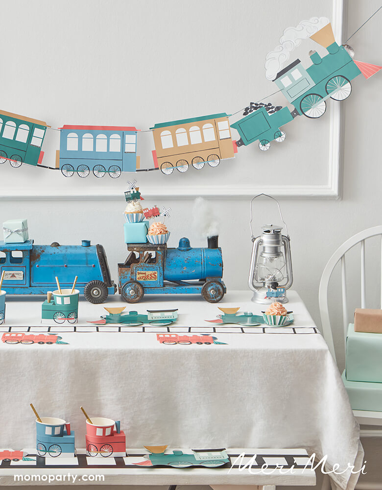 An adorable party decoration set up for kid's train themed party with Meri Meri's train garland on hung on the wall, and a table with a vintage toy train as the centerpiece, carrying party cupcakes dressed with Meri Meri's train cupcake kit, on the table and the bench train tracks was drawn and Meri Meri's train shaped plates, napkins and party cups were placed along side the train track - a perfect inspiration for kid's train themed celebration or a chugga chugga two two themed 2nd birthday party.