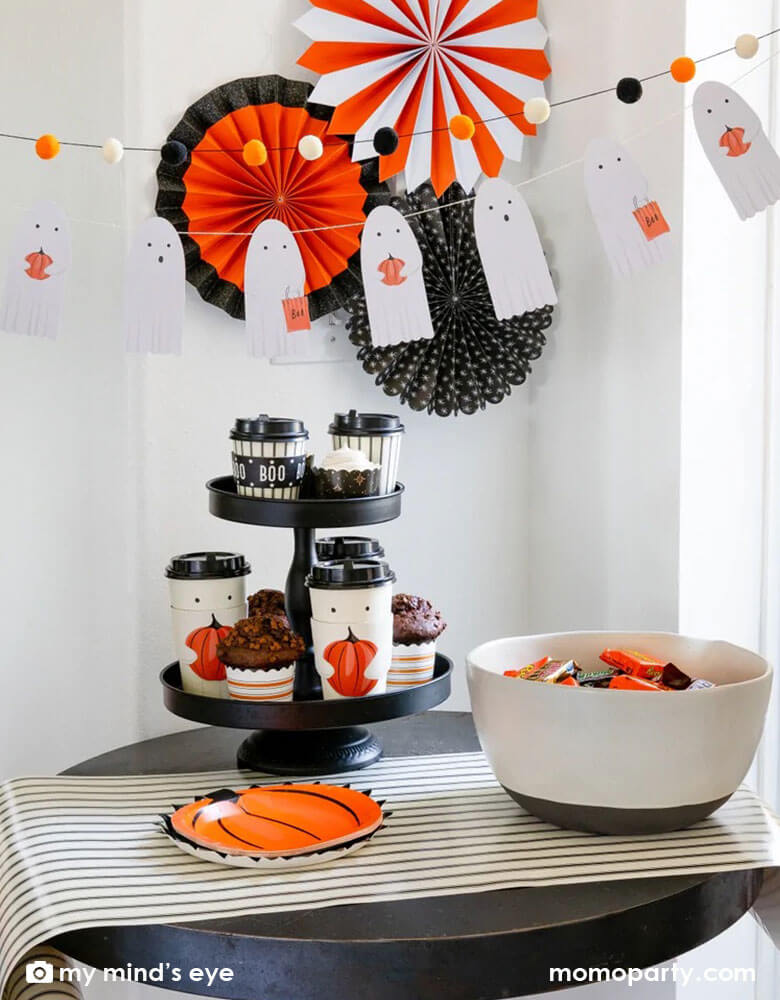 A spooky yet adorable mini table set up next to the window featuring My Mind's Eye boo crew ghost banners with black and orange pom poms, with vintage Halloween paper fans in the back, on the table were a bowl of Halloween candies and to-go cups in ghost design, ready for a fun trick or treat Halloween night!