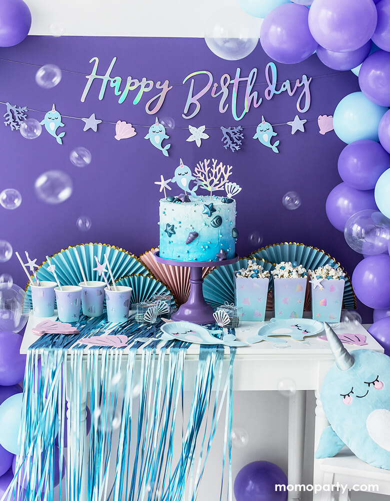 Under The Sea Narwhal Party Table Idea, with a iridescent happy birthday garland and Narwhal shell garland, and purple, light blue, and clear latex balloons mixed balloon garland as decoration. A ombre cake with Narwhal cake topper on the purple cake stand, Under The Sea Ombre Cups with paper party straws on top of blue Party curtain,  Narwhal paper plates, Pink Shell Napkins, Under The Sea Popcorn Treat Boxes with popcorns, iridescent confettis around on the table for a dreamy under the sea birthday party