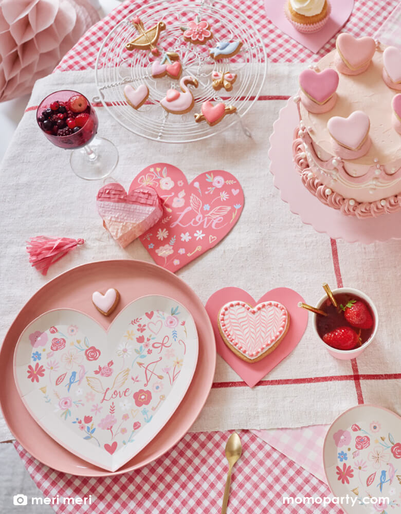 A Valentine's Day party table with pink buttercream cake decorated with heart shaped macarons in beautiful shades of pink, along with Meri Meri's 9.25 x 8.5 inch VALENTINE HEART DIE CUT PLATES paired with the VALENTINE HEART DIE CUT napkins in red with beautiful illustrations of love, flowers, doves and hearts, plus strawberry infused drinks in Meri Meri pink tone party cups and heart shaped piñata spread across the table, makes great party inspirations and decoration ideas for Valentine's Day celebration!