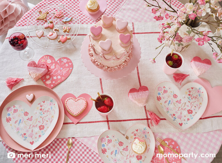 A Valentine's Day party table with pink buttercream cake decorated with heart shaped macarons and adorable Valentine's Day sugar cookies in the shape of hearts, swans and doves, along with Meri Meri's Valentine Heart die cut plates paired with the Valentine's Heart napkins in red with beautiful illustrations of love, flowers, doves and hearts, plus strawberry infused drinks in Meri Meri pink tone party cups and heart shaped piñata spread across the table.