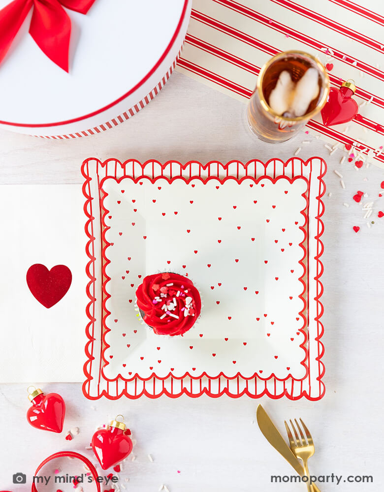 A sweet Valentine's Day celebration table filled with Momo Party's 8" square scattered red heart scallop plate by My Mind's Eye, Valentine Red Foil Heart Guest Napkins, heart shaped decoration, confetti, drinks and gold utensils over a Believe Christmas Red Striped Table Runner, for a sweet valentine's day party