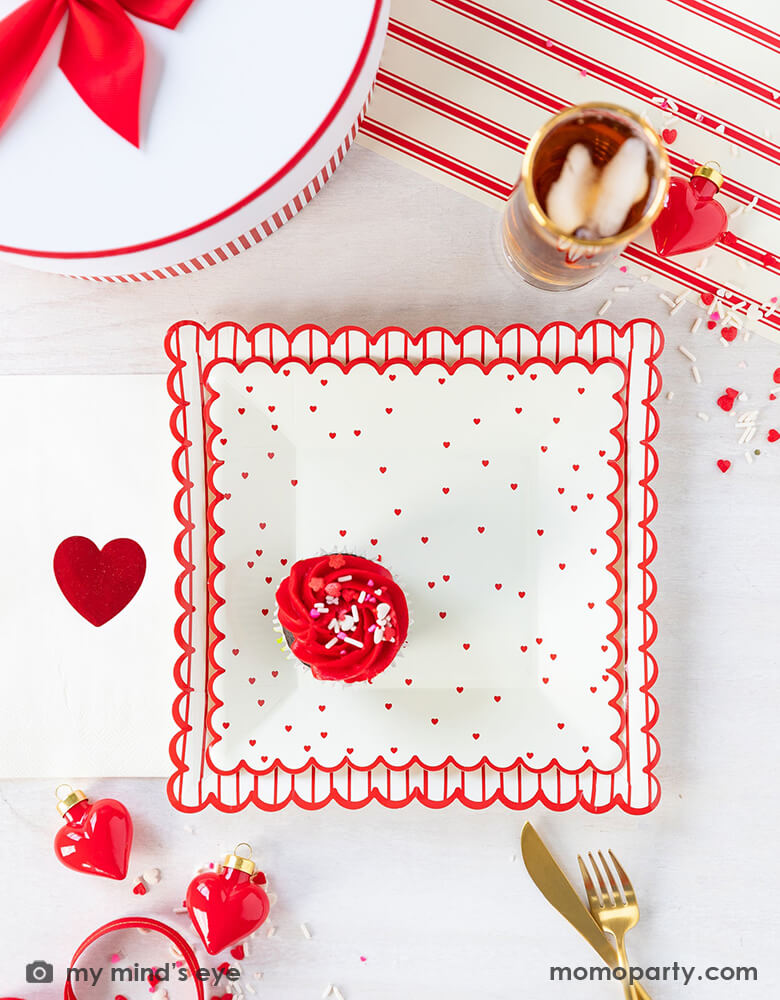  A sweet Valentine's Day celebration table filled with Momo Party's 8" square scattered red heart scallop plate by My Mind's Eye, Valentine Red Foil Heart Guest Napkins, heart shaped decoration, confetti, drinks and gold utensils over a Believe Christmas Red Striped Table Runner, for a sweet valentine's day party